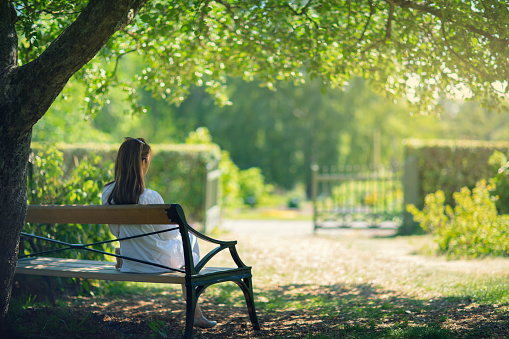 A woman sitting down on a bench in a garden in the shade of a tree and enjoying the surroundings.