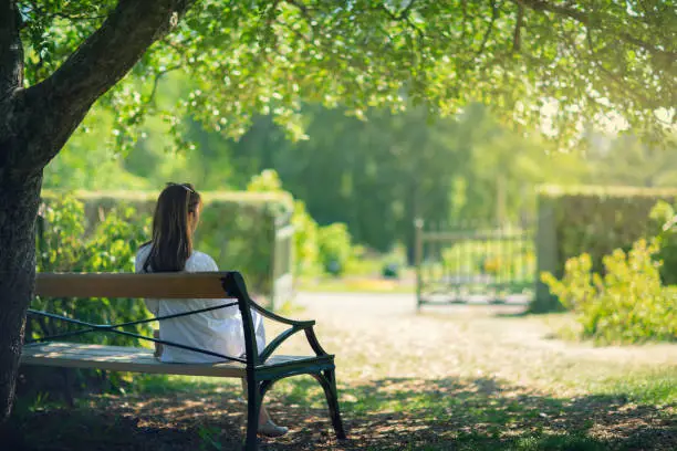 A woman sitting down on a bench in a garden in the shade of a tree and enjoying the surroundings.
