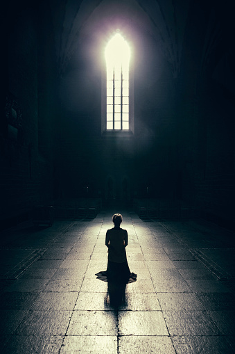 A woman kneels down in a prayer on a church floor in the light of a single window.