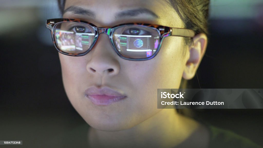 Population data screen Close up stock image of a young asian woman staring at a computer screen which is reflected in her glasses. The reflection depicts graphs; people & data. Eyeglasses Stock Photo