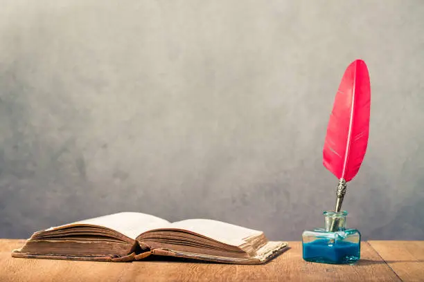 Photo of Vintage old red quill pen with inkwell and open book on wooden table front concrete wall background. Retro style filtered photo