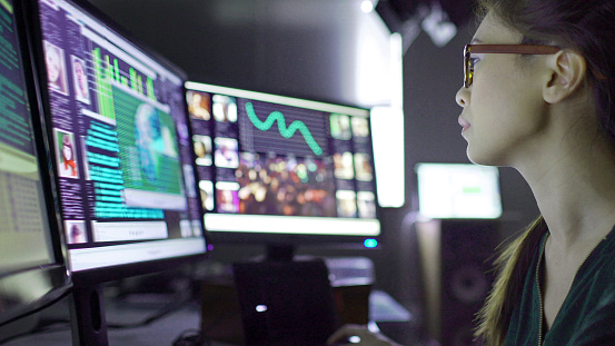 Close up stock image of a young asian woman sitting down at her desk where she’s surrounded by 3 large computer monitors displaying out of focus images of people as thumbnails; crowds; graphs & scrolling text.