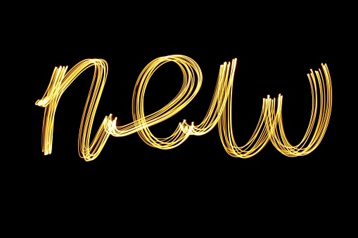 Long exposure photograph of light painted letters, spelling the word 'new' in joined up lettering, made with gold fairy lights, against a black background. Happy new year series concept.