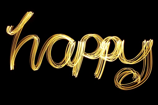 Long exposure photograph of light painted letters, spelling the word 'happy' in joined up lettering, made with gold fairy lights, against a black background. Happy new year series concept.