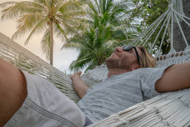 istock Young man relaxing on hammock in tropical climate enjoying serene environment with palm trees and green grass garden. People travel relax concept in vacations 1054722430