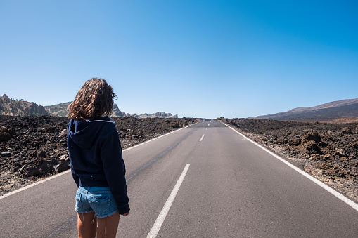 travel and destination concept with long asphalt road in the middle of the mountains and cute girl viewed from rear looking to the end of the line. traveler and wanderlust image with beautiful girl.