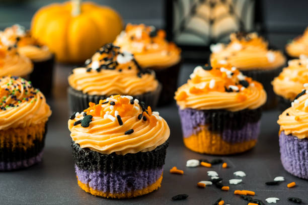 Halloween Cupcakes with Orange Frosting Halloween cupcakes with purple, black and orange cake bottom frosted with a swirled orange frosting. Cupcakes are decorated with Halloween themed sprinkles, bats , ghosts and multi colored sprinkles. A pumpkin and spider web lantern can be seen in the background. halloween cupcake stock pictures, royalty-free photos & images