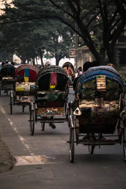 Perfectly lined up and beautifully colored rickshaws or tuktuks driving down a tree lined street in Dhaka, Bangladesh