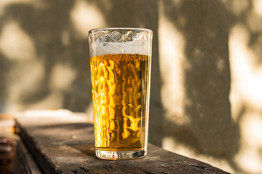 A glass of beer with alcohol. Golden, intoxicating drink in a transparent mug, stands on a wooden stand in the rays of daylight. Top white and airy foam.