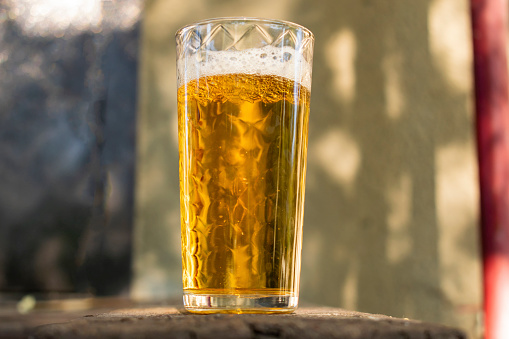 A glass of beer with alcohol. Golden, intoxicating drink in a transparent mug, stands on a wooden stand in the rays of daylight. Top white and airy foam.