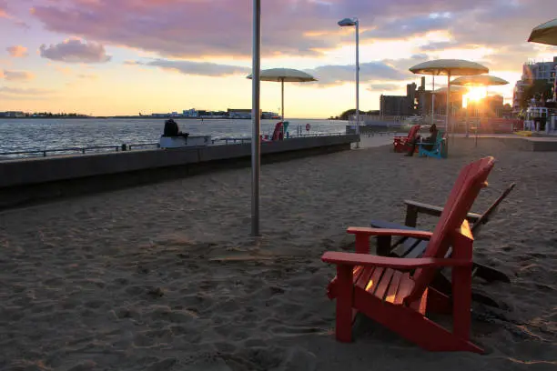 An almost empty Toronto HTO Park at sunset. With several Muskoka armchairs and sun umbrellas on the artificial beach. The sun was close to the horizon. The sun rays illuminated the fluffy clouds on the sky.