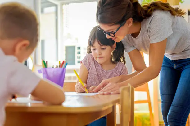 Photo of Children drawing with smiling preschool teacher assisting them