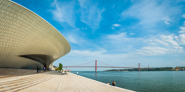 The iconic ceramic tiles of the MAAT Museum of Architecture, Art and Technology overlooking tourists on the blue waterfront of the River Tagus beneath the 25 de Abril Bridge in the heart of Lisbon, Portugal’s vibrant capital city.