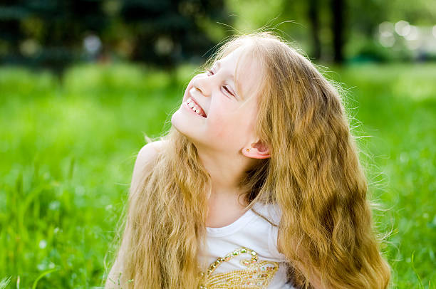 Toothless portrait of beautiful little girl with smiling eyes and long brunette wavy hair looking to camera on green grass background. Kid happy face TOP VIEW SHOT