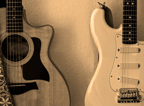 Close frame in sepia tone of part of the body of an acoustic guitar showing sound hole, bridge and part of neck next to part of the body of a white electric guitar showing pickups, bridge and part of neck