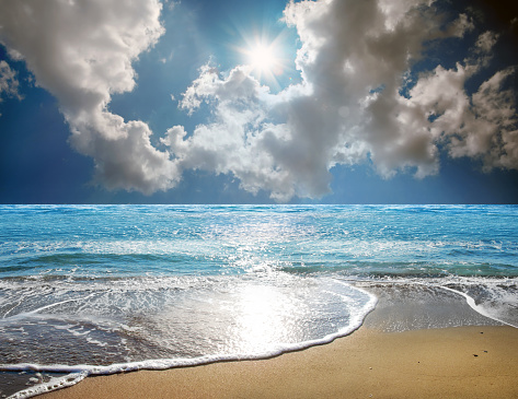 Seascape image of sandy beach and waves on shiny sea over sunny and cloudy sky