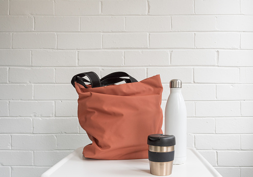 Reusable coffee cup, insulated drink bottle and shopping tote bag on white table against brick wall with copy space