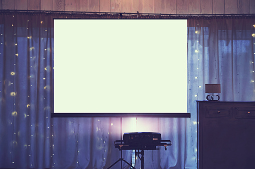 Front view of decorated wedding room with empty white projector screen in the centre. Equipment for video and slideshow projection at a festive event. Banquette hall with a board for projection