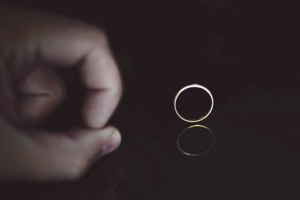 close up of man's fingers on the reflecting black surface pushing on the wedding ring at the black background. male hand on the black table near the gold ring set edgewise. man put ring off. divorce. - unwillingness imagens e fotografias de stock