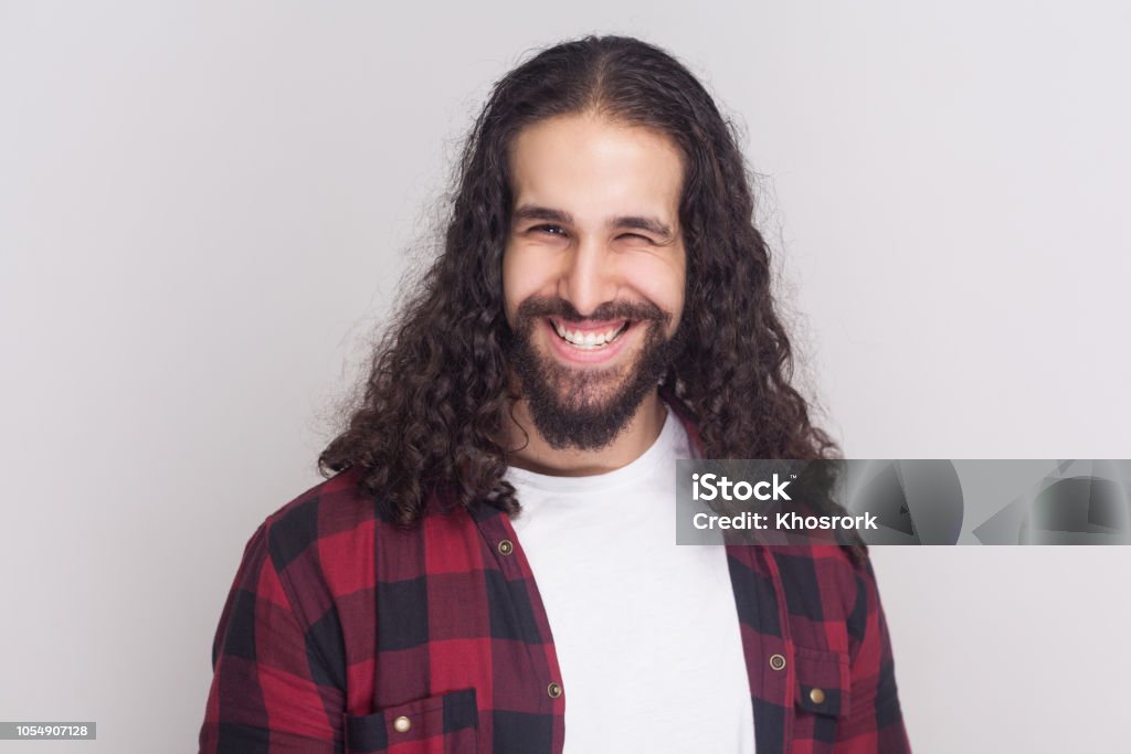 Funny Handsome Man With Beard And Black Long Curly Hair In Casual Style Checkered Red Shirt Standing Winking And Looking At Camera With Toothy Smile Stock Photo - Download Image Now - iStock Funny Handsome Man With Beard And Black Long Curly Hair In Casual Style Checkered Red Shirt Standing Winking And Looking At Camera With Toothy Smile Stock Photo - Download Image Now - iStock