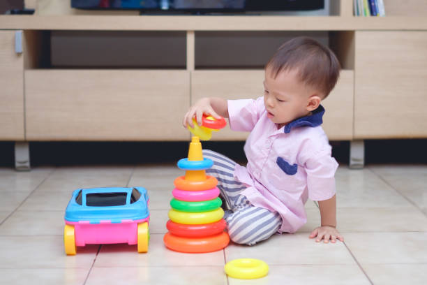 Cute little Asian 18 months / 1 year old toddler boy play with educational colorful plastic pyramid toy / stacking ring toy in living room stock photo