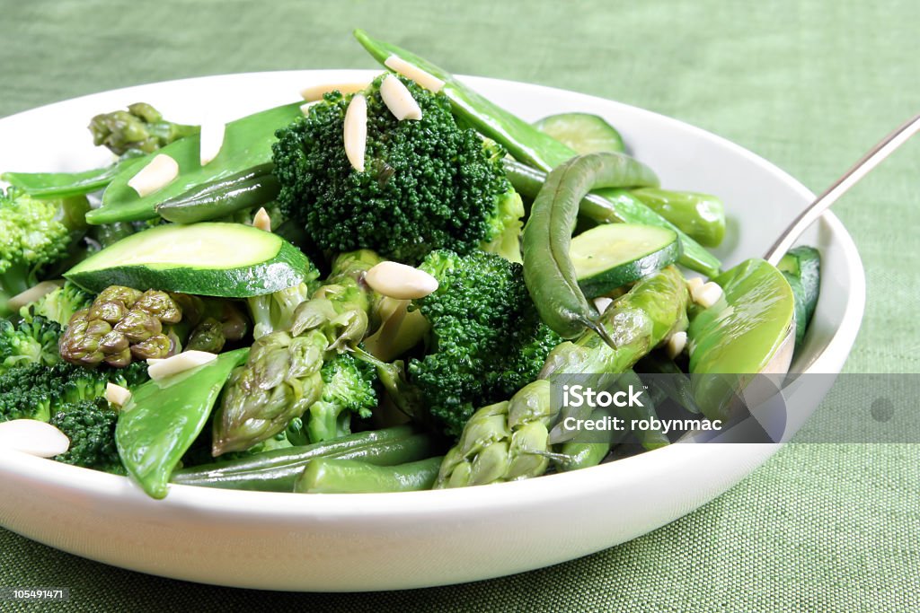 A bowl of broccoli, mangetout and asparagus with a spoon Serving bowl of mixed green vegetables, topped with toasted almond.  Delicious healthy eating.  Please see also: Almond Stock Photo