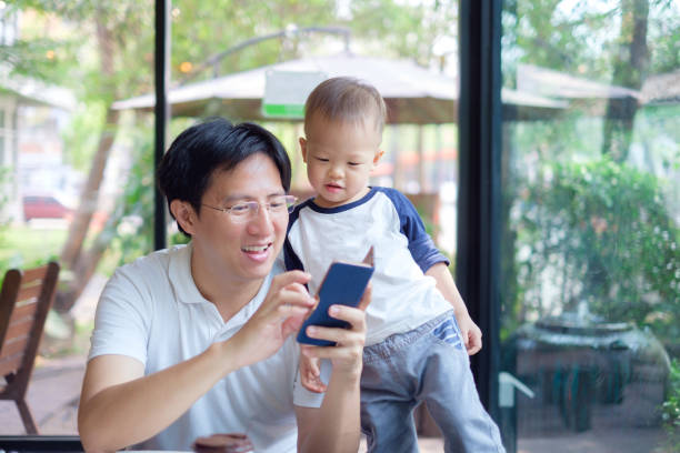 азиатский папа и сын, сидя в городском кафе смотреть в смартфоне, отец и милый маленький 1 год / 18 месяцев малыш мальчик ребенок проводит врем - child looking messy urban scene стоковые фото и изображения