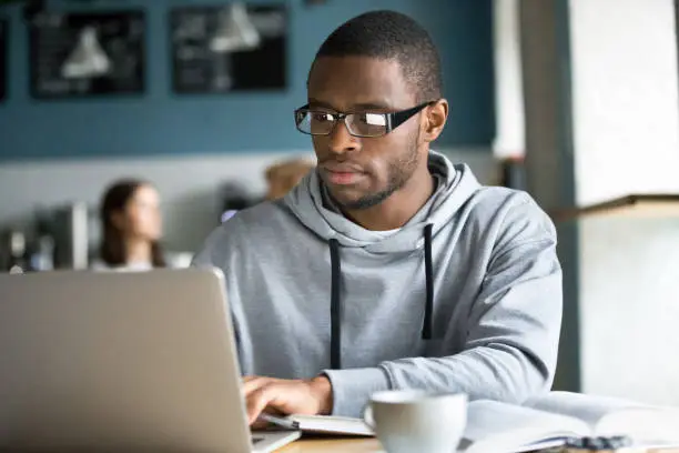 Serious black guy in glasses work at laptop sitting in coffeeshop, concentrated African American student study online at computer having coffee in near cafÃ©, focused afro use gadget browsing internet