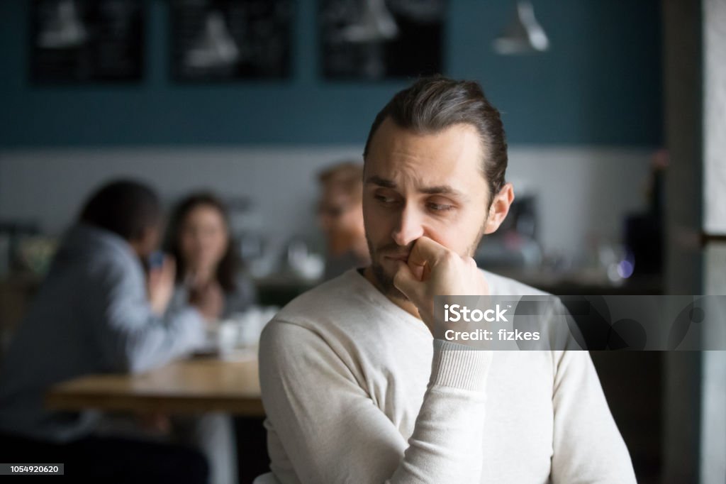 Upset male outcast feel lonely sitting alone in cafe Upset millennial outsider feel offended lack company, young outcast guy suffer from discrimination, jealous of friends hang out together in cafÃ©, envious male loner depressed sit alone in coffeeshop Men Stock Photo