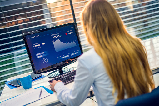 Rear view of an unrecognizable female financial investment fund manager using her office computer to analyze a cryptocurrency graph.