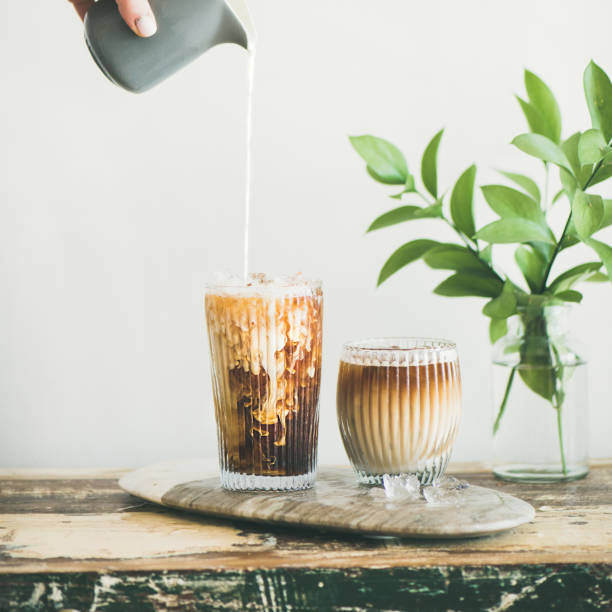 Iced coffee in tall glass with cream, square crop Iced coffee in tall glasses with milk poured over from pitcher by hand, white wall and green plant branches at background, copy space, square crop. Summer refreshing beverage ice coffee concept cafe macchiato stock pictures, royalty-free photos & images