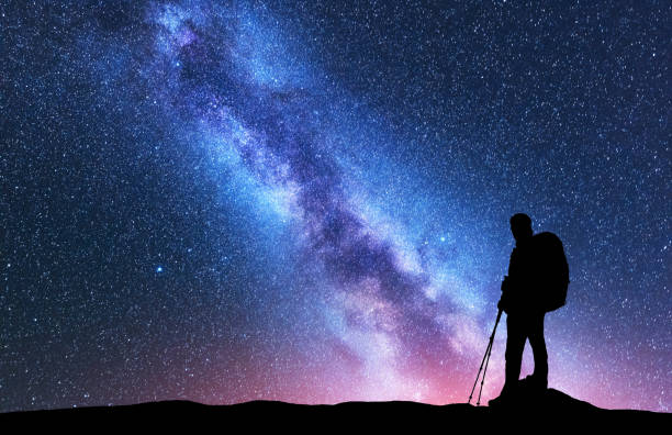 silhouette of man with backpack and trekking poles against amazing purple milky way at night. space. landscape with man, bright milky way, sky with stars. beautiful galaxy. travel. starry sky. nature - milky way galaxy space star imagens e fotografias de stock