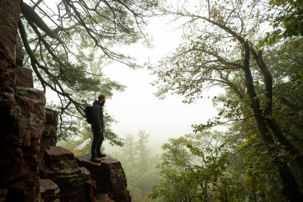active man hiking through scenic dense foggy forest overlooking steep mountain cliff with rocks and dramatic trees with backpack on running trail. devils lake wisconsin - devils lake imagens e fotografias de stock