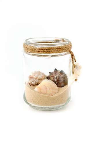 Seashells and sand in a handmade glass jar decorated with mini seashells and isolated on white background. Low angle view.