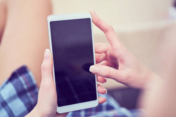 istock Back view of a woman hands using smart phone with blank screen close up. White phone with a black screen in the hands of a young girl lying on the floor with her feet on the couch 1054952124