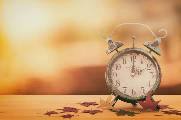 Photo of Image of autumn Time Change. Fall back concept. Dry leaves and vintage alarm Clock on rustic wooden table.