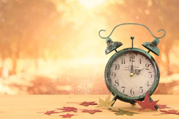 Photo of Image of autumn Time Change. Fall back concept. Dry leaves and vintage alarm Clock on rustic wooden table.