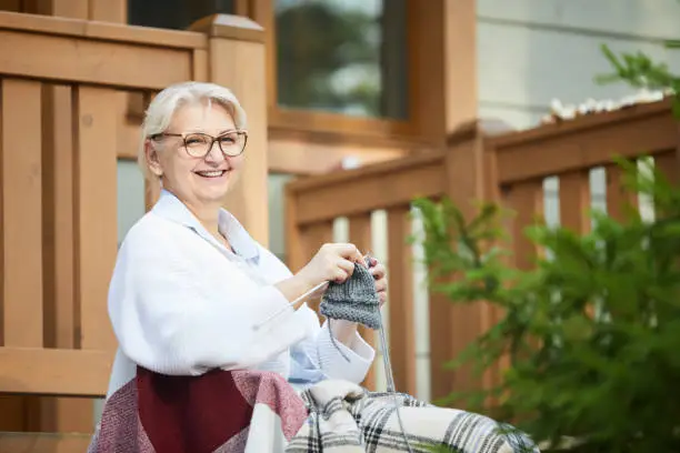 Cheerful grandma siting on rocking chair by terrace, looking at you and knitting warm woolen scarf