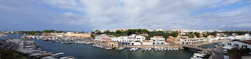 A wide panorama view of the town of ciutadella in menorca showing boats moored along the canal and surrounding buildings in bright summer sunlight
