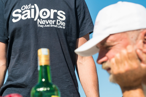 Portrait of Senior man with daisy eyes looking at brandy bottle and is thinking to have a slip or two more while sitting on sailing boat on the open Adriatic Sea in Croatia. In background is on other man t-shirt sign 
