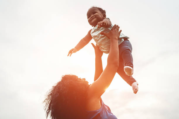 felice madre africana che gioca con sua figlia all'aperto - mamma e bambino afro che si divertono insieme - concetto di famiglia, felicità e amore - morbida attenzione al viso della donna - baby mother summer park foto e immagini stock