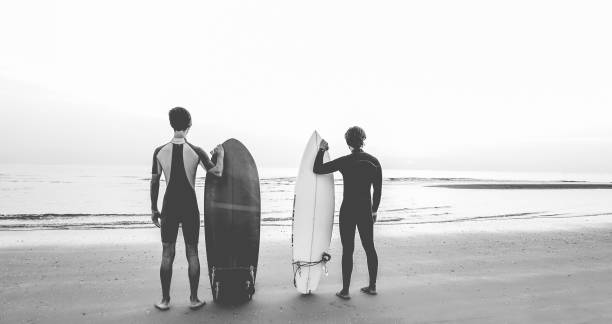 junge surfer warten die wellen am strand - sport-freunde, die immer bereit zum surfen - extreme sport, lifestyle und erholung jugendkonzept - schwarz / weiß bearbeiten - surfen fotos stock-fotos und bilder