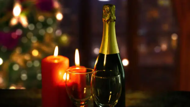 Close up of red burning candles with two glasses and a bottle of champagne in blurred Christmas tree background