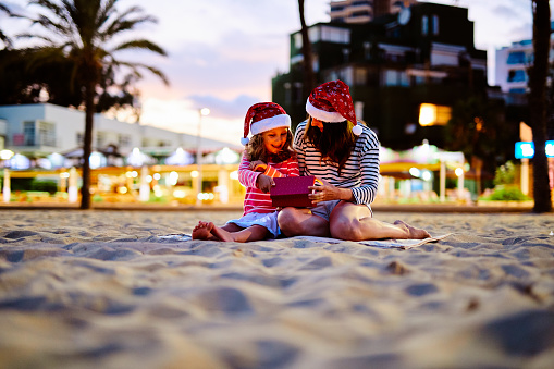 Mother giving christmas present to her daughter on the beach, at night, both wearing santa hats
