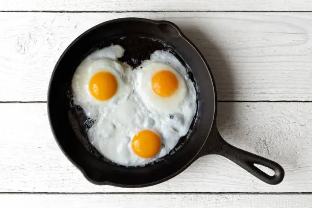 Three fried eggs in cast iron frying pan isolated on white painted wood from above.