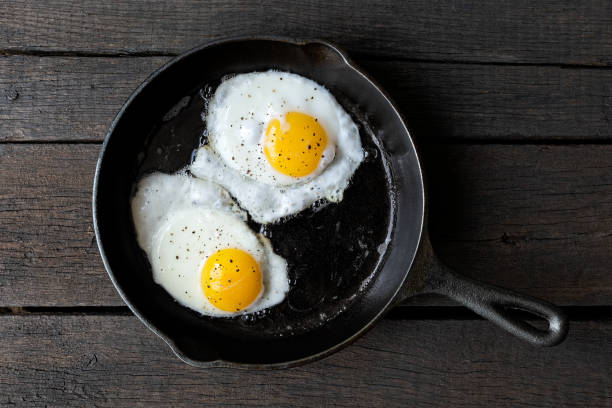 istock Two fried eggs in cast iron frying pan sprinkled with ground black pepper. Isolated on dark painted wood from above. 1055033468