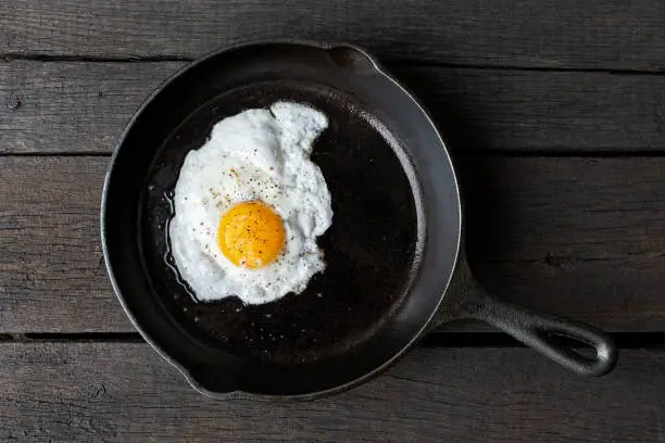Single fried egg in cast iron frying pan sprinkled with ground black pepper. Isolated on dark painted wood from above.