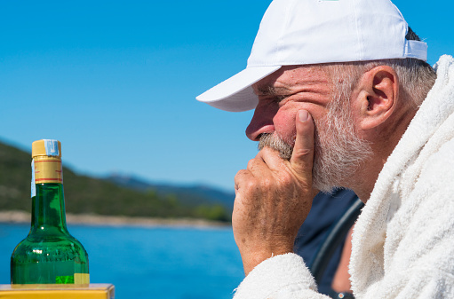 Portrait of Senior man thinking hard, with daisy eyes looking at brandy bottle and is thinking to have a slip or two more while sitting on sailing boat on the open Adriatic Sea in Croatia with Long Island (Dugi otk) in background. .