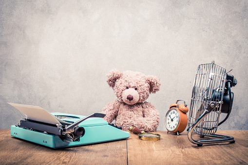 Teddy Bear toy sitting at the old wooden desk with mint green retro typewriter, orange alarm clock and cooling fan front concrete wall background. Vintage style filtered photo