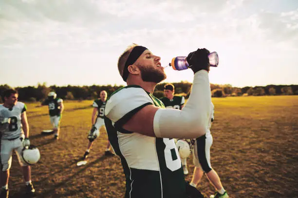 American football player drinking water from a bottle while standing on a field during a team practice